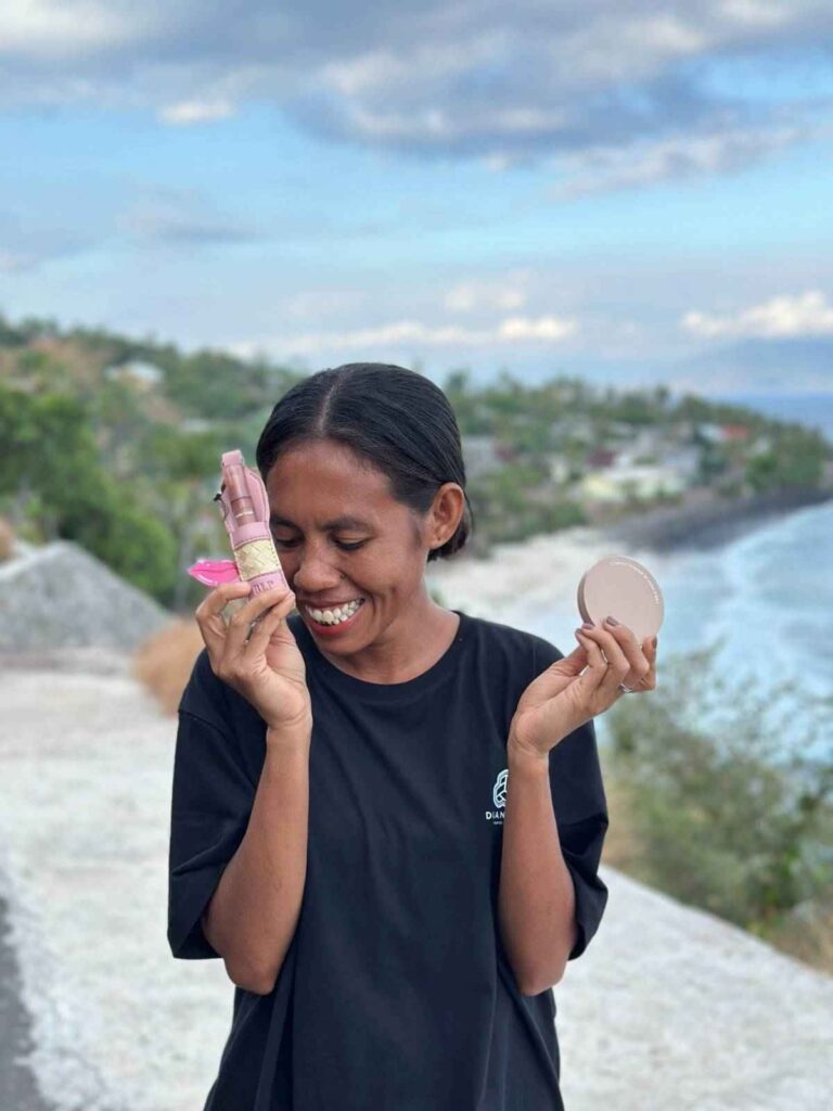 Seorang wanita  tersenyum memperlihatkan lipstick sleeve anyaman dengan compact powder dengan latar Flores NTT